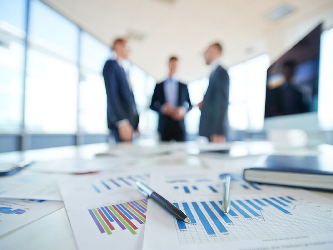 Im Vordergrund liegen Stifte und Papiere auf einem Schreibtisch; im Hintergrund sind verschwommen drei Mitarbeiter zu sehen.