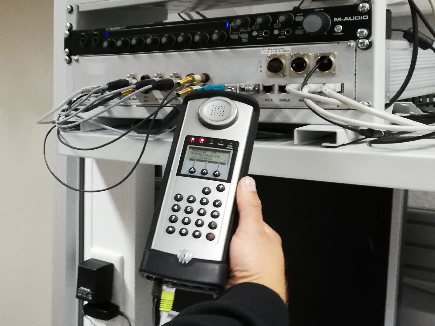 Eine Hand hält ein Messgerät, das an einen im Hintergrund sichtbaren Kasten mit Kabeln angeschlossen ist.