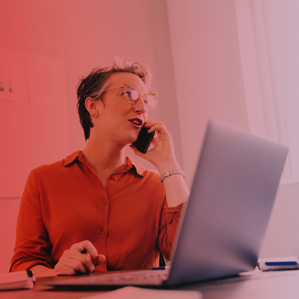 Eine Frau mit Brille und rotem Hemd telefoniert mit einem Avaya Aura Telefon und arbeitet an einem Laptop in einem Büro.